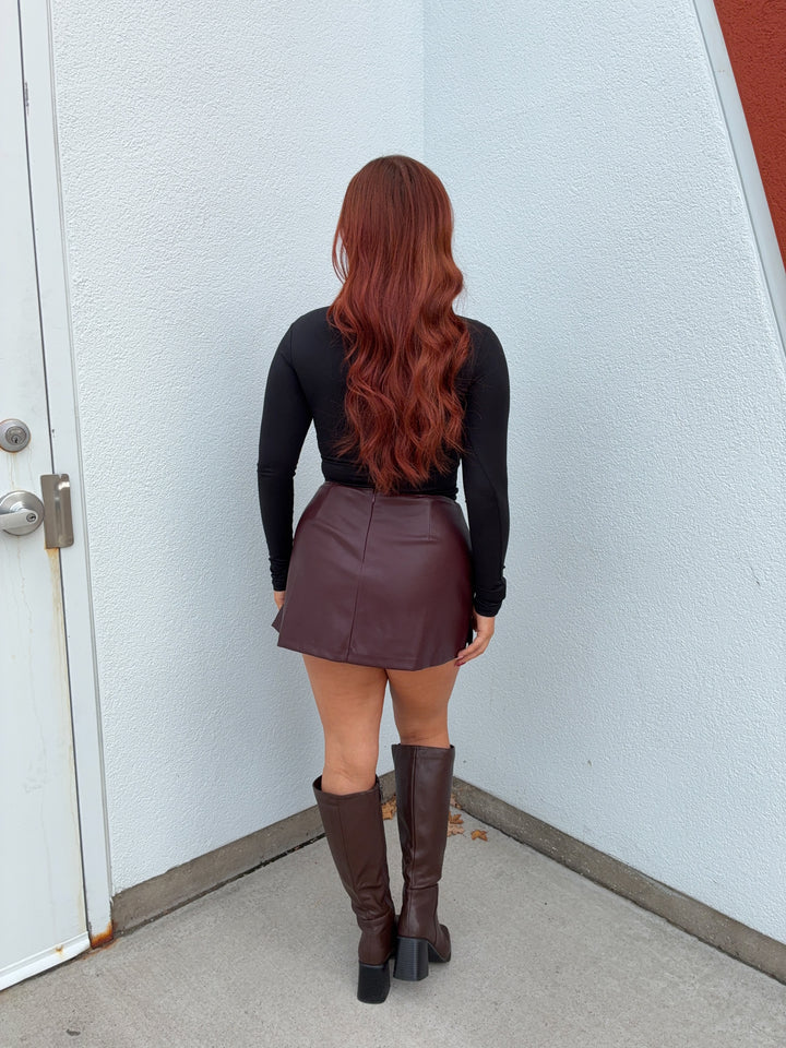 Person wearing a black long-sleeve top and brown leather skirt with knee-high boots, standing against a light blue wall.