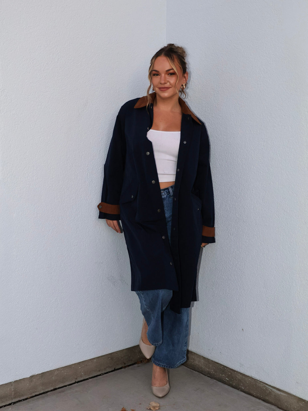 Woman wearing a navy coat, white top, and blue jeans against a white wall.