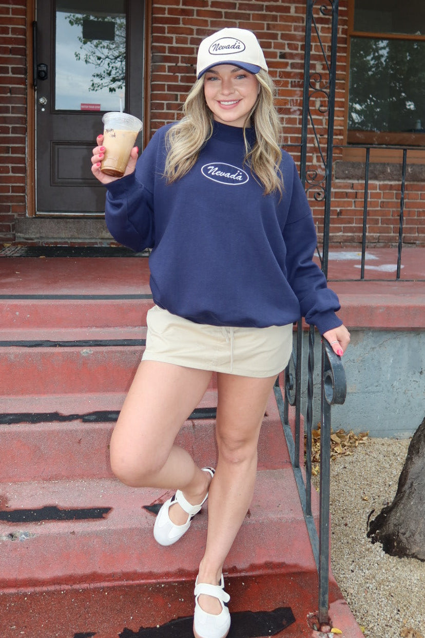 A woman posing on the red brick steps of a building wearing a navy blue sweatshirt with a white 'Nevada' logo, a white baseball cap, beige shorts, and white shoes, while holding an iced coffee.