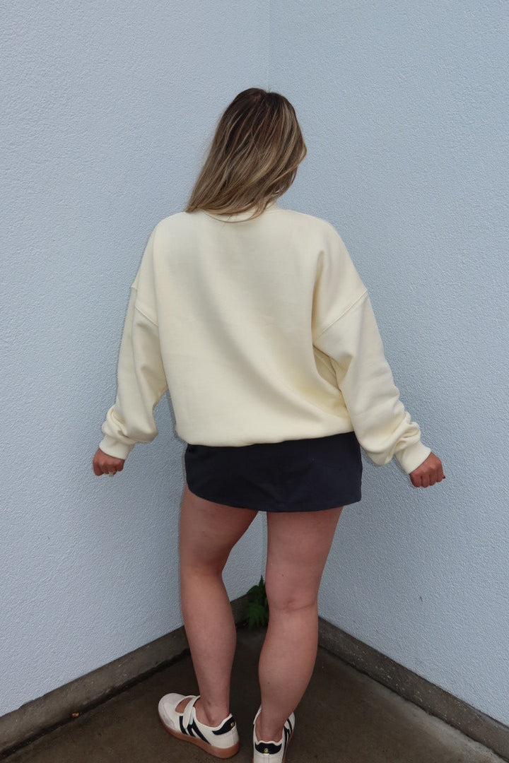 A woman viewed from behind wearing a cream-colored crewneck sweatshirt, a dark navy mini skirt, and white sneakers with black stripes, standing in the corner of a building with a light blue wall and a red accent panel.