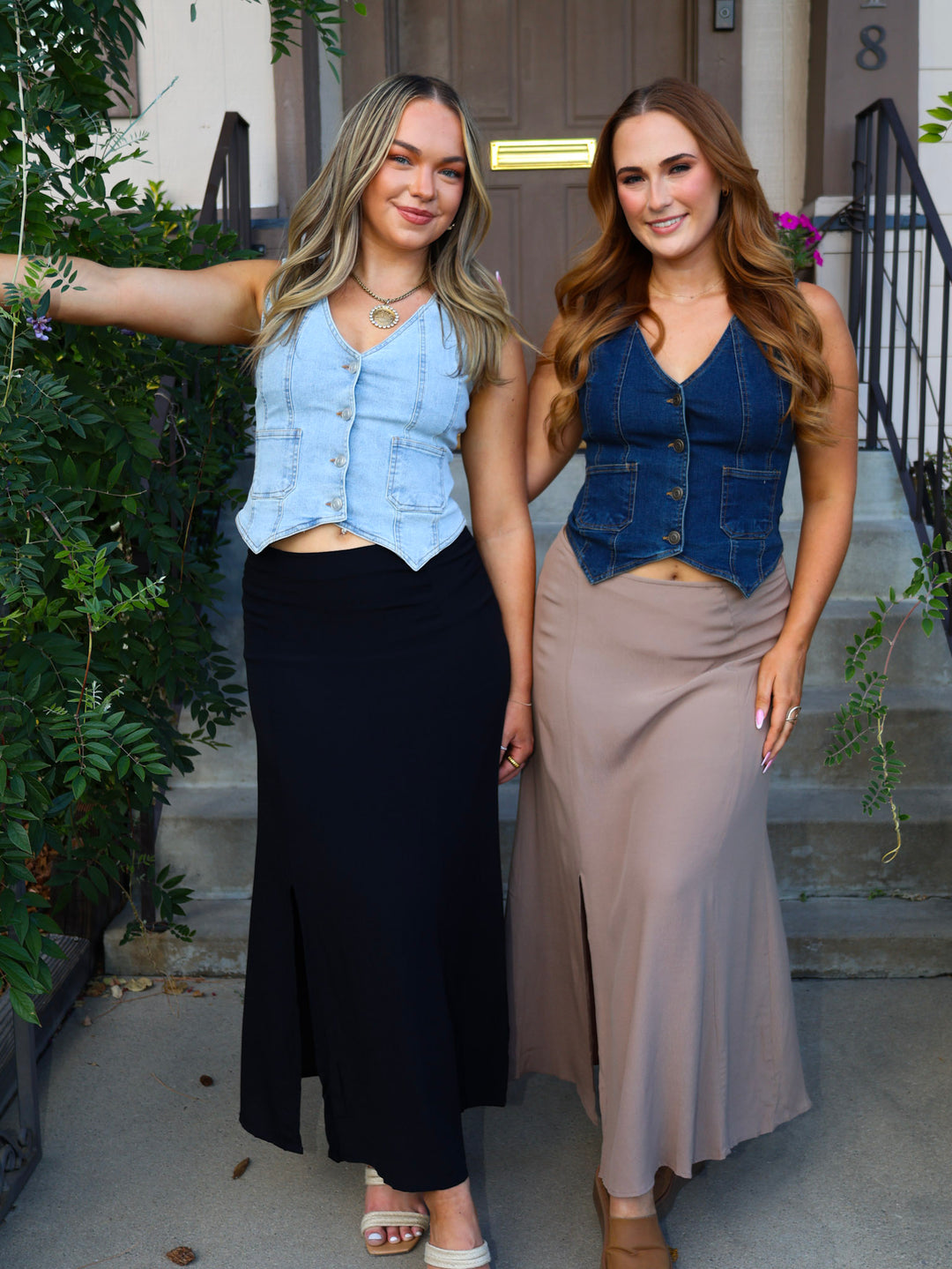 Two women standing outdoors, wearing matching denim vests and long skirts.