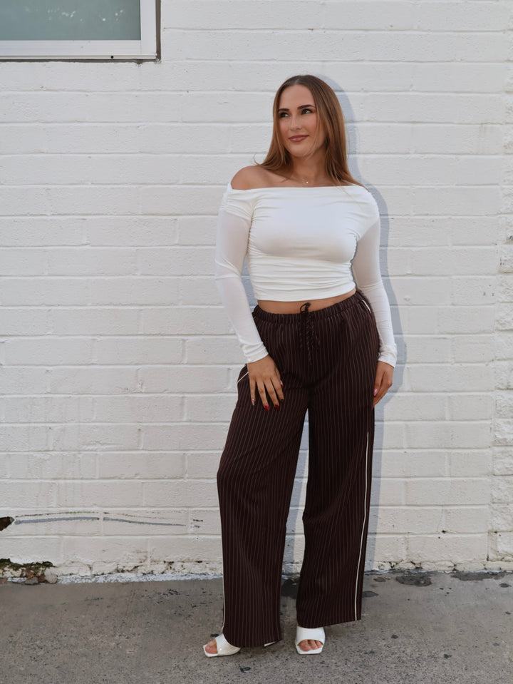 Woman wearing a white off-shoulder top and dark pinstripe pants against a white brick wall.