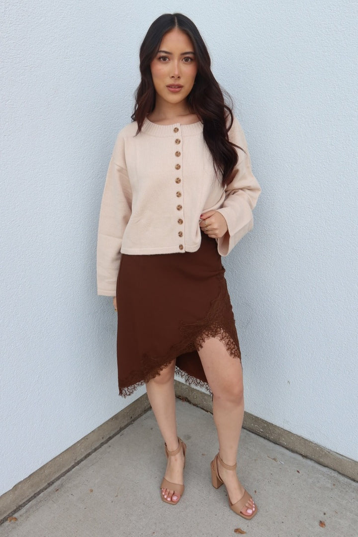 Model wearing the brown Lucy asymmetric lace trim satin midi skirt with a cream button cardigan and tan heeled sandals