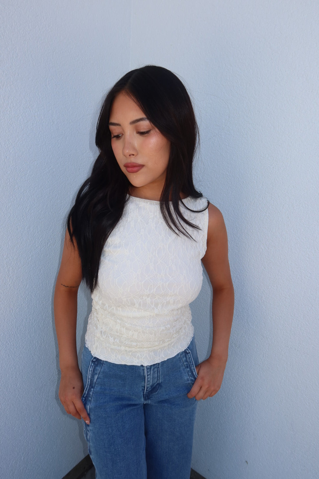 A woman with long black hair wearing a textured white sleeveless top and blue jeans, standing against a light blue wall.