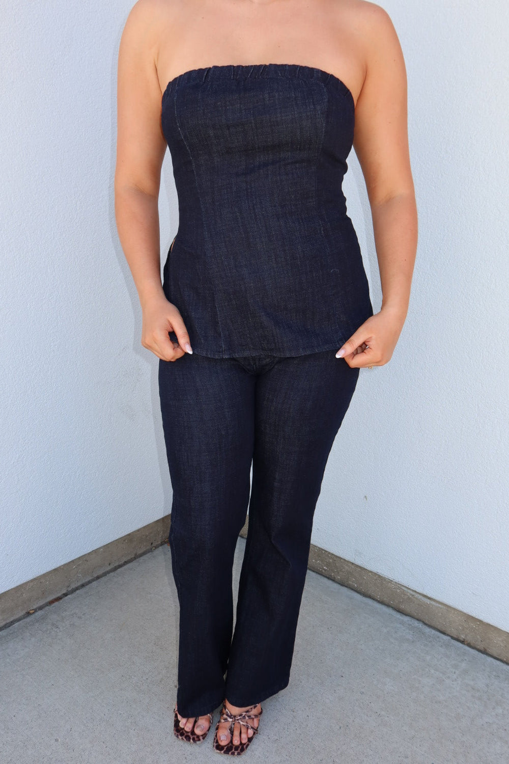 A woman modeling a dark indigo denim strapless tube top and matching dark blue denim flare jeans, standing against a plain light gray wall.