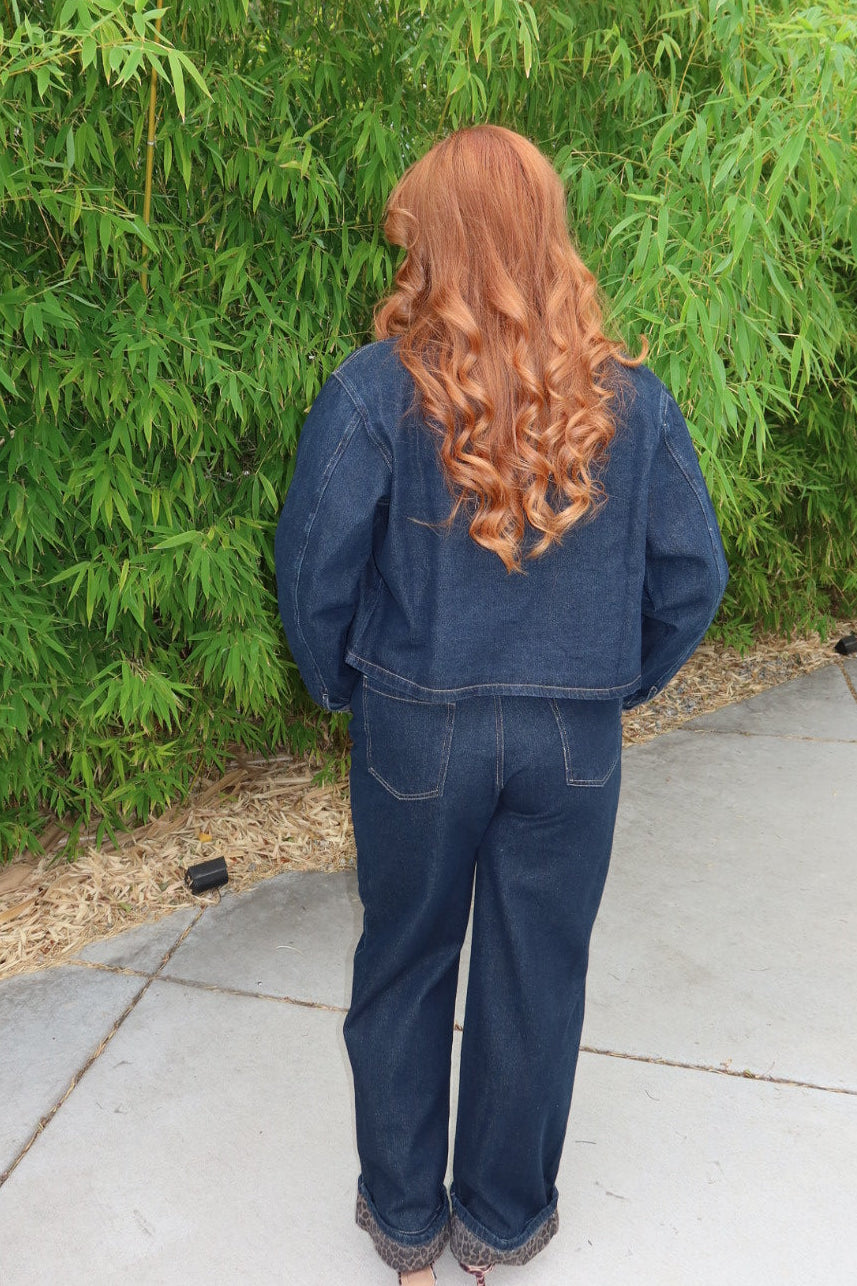 A person with long, wavy red hair seen from behind, wearing a dark blue denim jacket and matching wide-leg denim pants, standing on a concrete path in front of a dense green bamboo hedge.