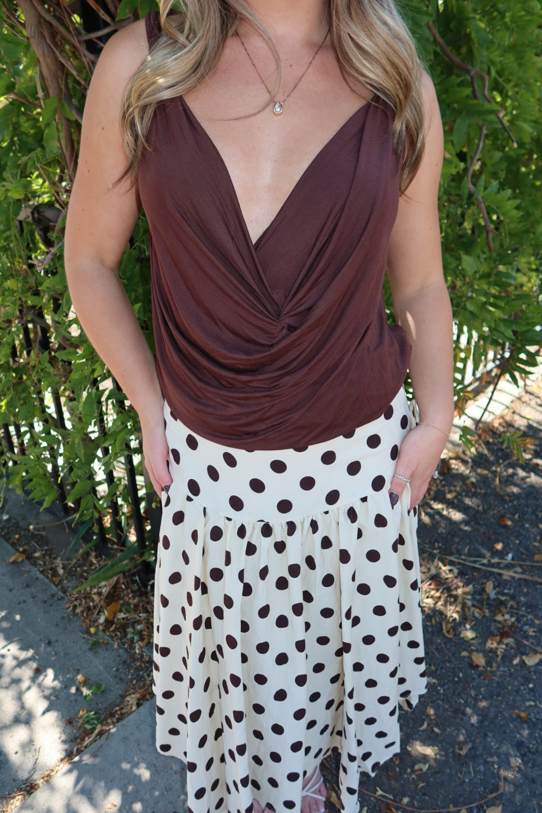 A woman wearing a brown draped wrap-style top and a white skirt with dark brown polka dots, standing outdoors in front of green foliage.