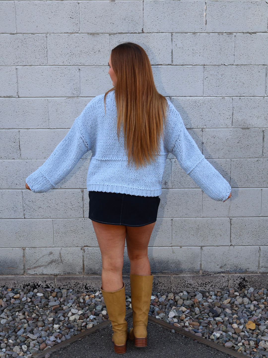 Person wearing a light blue sweater and black skirt with tan boots standing against a gray brick wall.