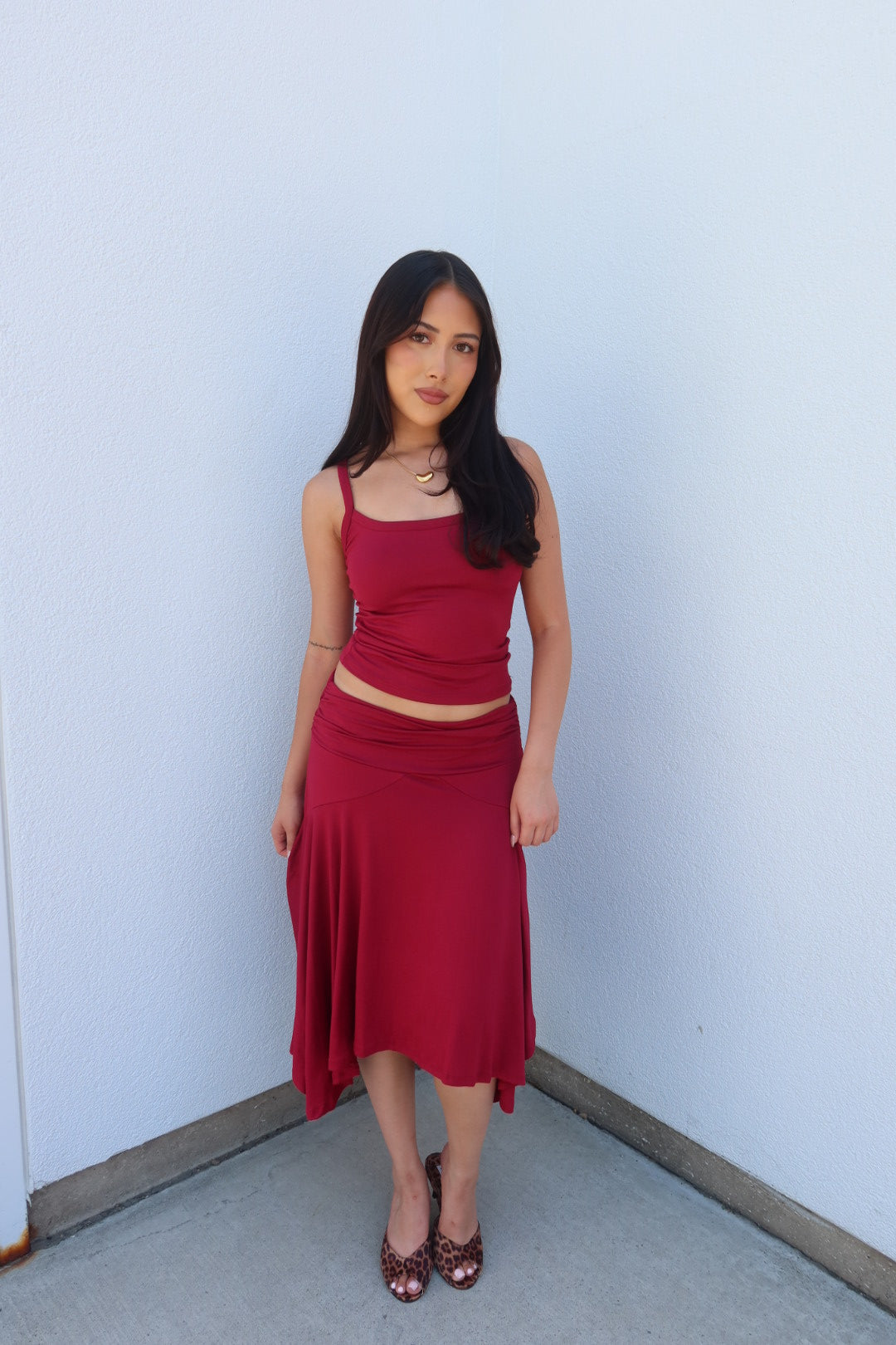A woman modeling a matching two-piece outfit consisting of a deep red spaghetti-strap crop top and a high-waisted, flowy midi skirt, paired with dark red heels, standing against a plain light blue-gray wall.