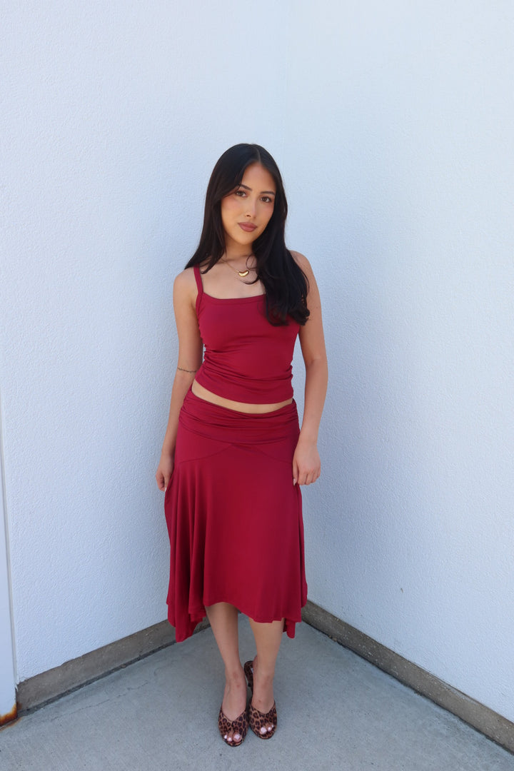 A woman modeling a matching two-piece outfit consisting of a deep red spaghetti-strap crop top and a high-waisted, flowy midi skirt, paired with dark red heels, standing against a plain light blue-gray wall.
