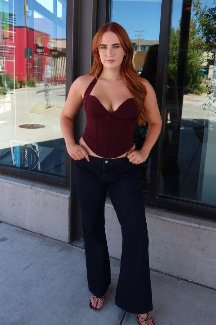 A woman with long red hair stands outdoors in front of a glass storefront, wearing a burgundy halter-style corset top and dark navy wide-leg jeans.