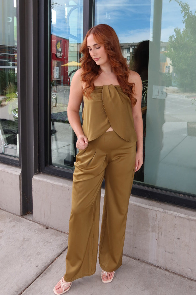 A woman with red hair wearing a mustard yellow strapless top and matching wide-leg pants, standing on a sidewalk in front of a building with large black-framed windows.