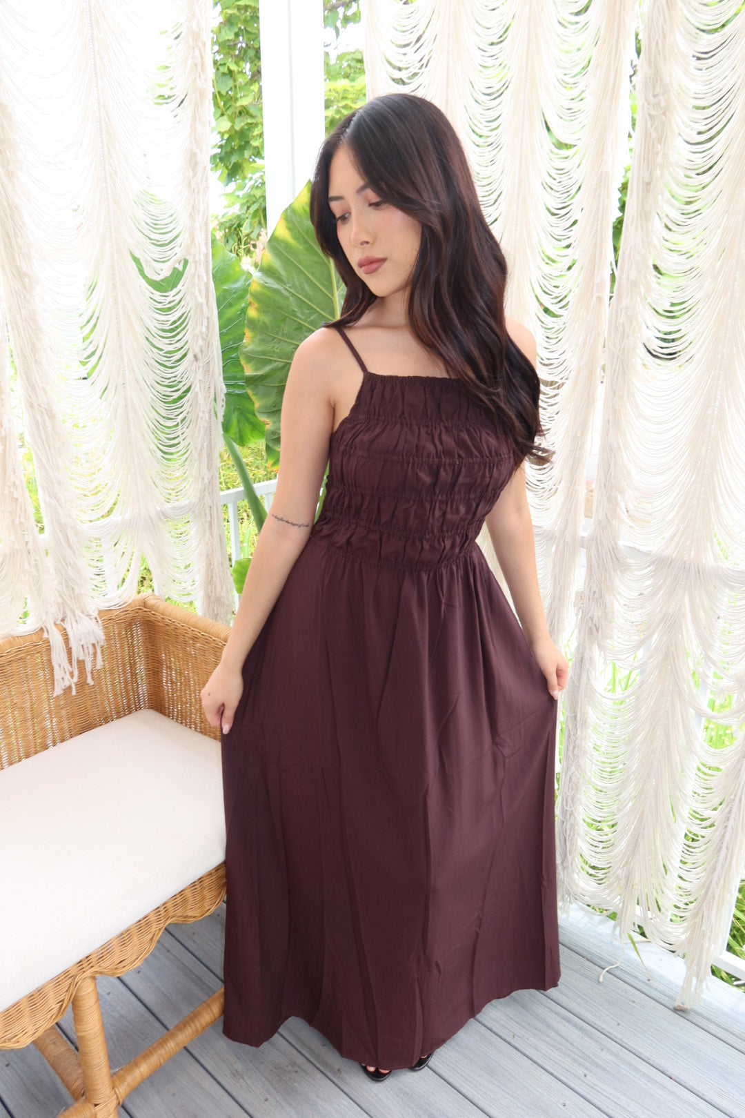A woman wearing a dark brown sleeveless maxi dress with thin straps and a smocked bodice, standing on a wooden deck in front of white macrame curtains.