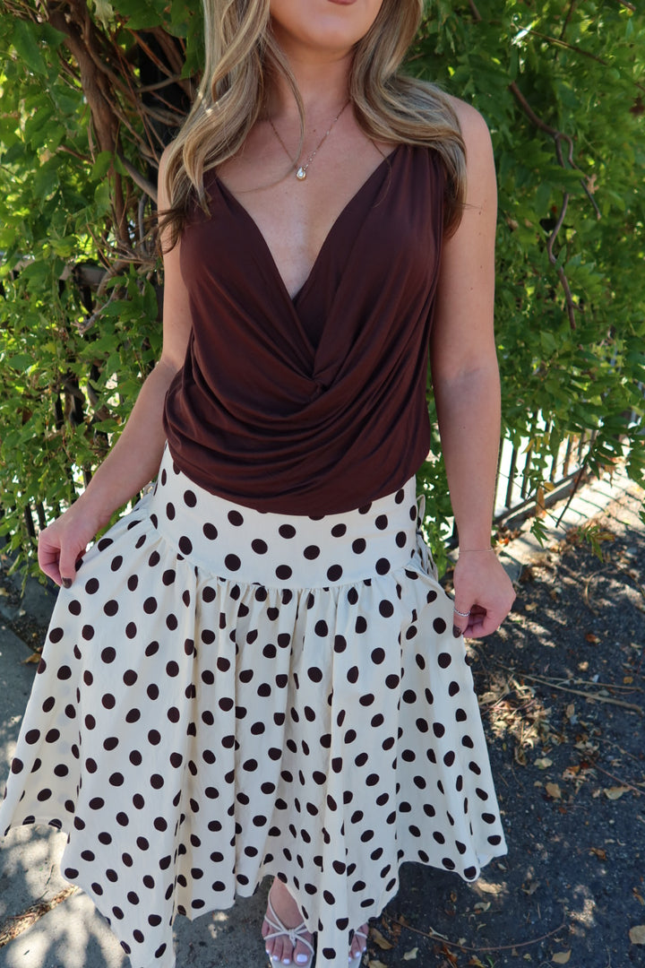 A woman wearing a brown draped sleeveless top and a white high-waisted skirt with dark brown polka dots, standing outdoors in front of green foliage.