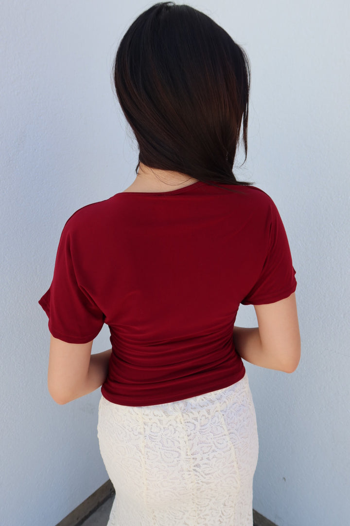 A woman viewed from behind wearing a fitted short-sleeve red top and a white textured skirt, standing against a plain light gray wall.