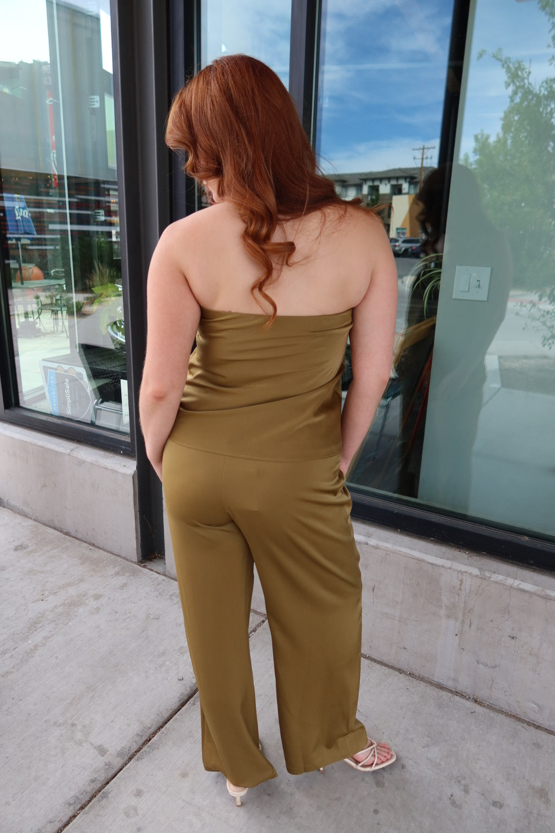 A woman with red hair is shown from behind wearing a strapless olive-brown jumpsuit while standing on a sidewalk in front of a glass storefront.