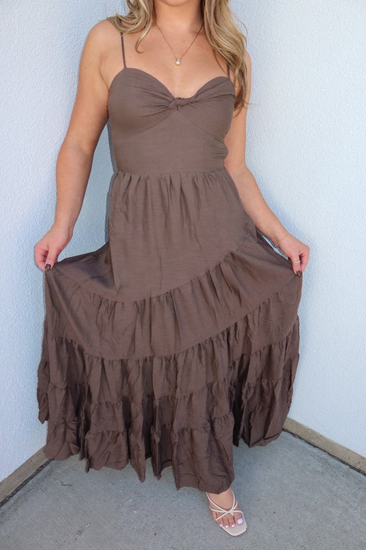 A woman wearing a brown tiered maxi dress with spaghetti straps and a sweetheart neckline, holding out the sides of the dress to show the ruffled layers, standing against a light blue wall.