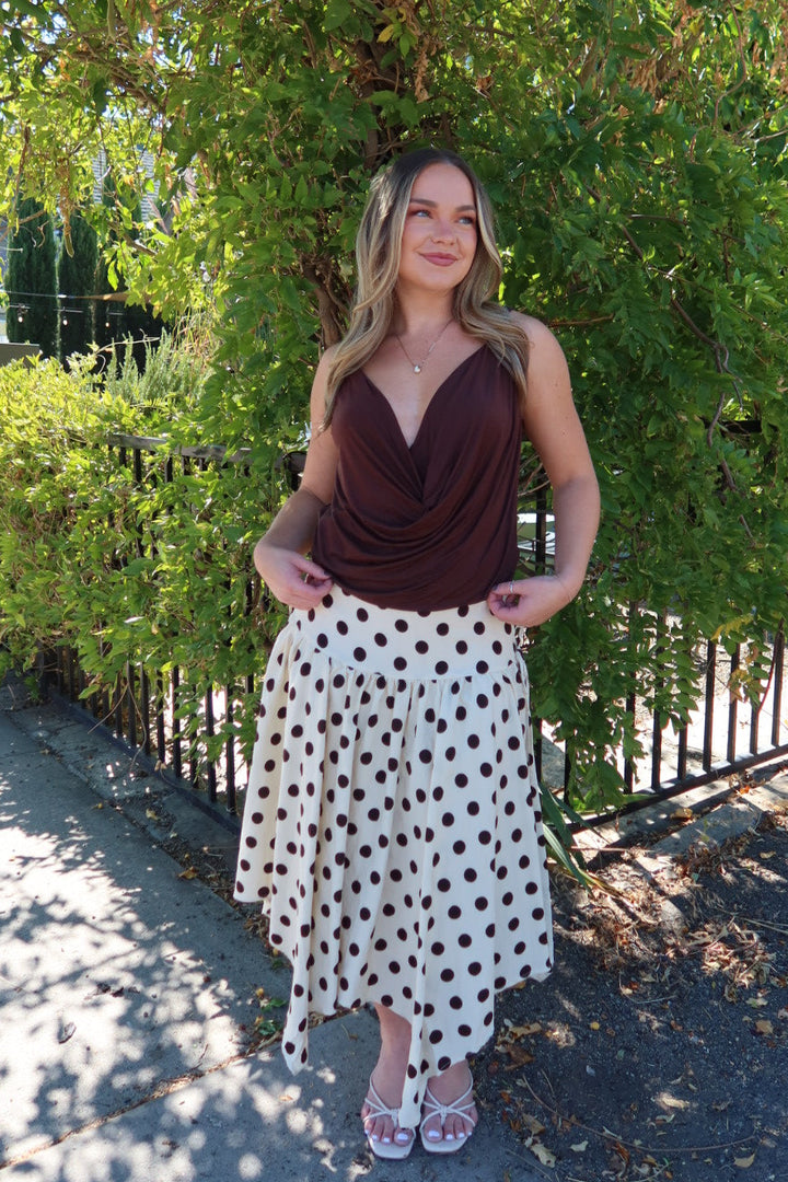 A woman stands outdoors in front of a leafy green tree, wearing a dark brown draped sleeveless top and a white midi skirt with black polka dots, paired with white strappy heels.
