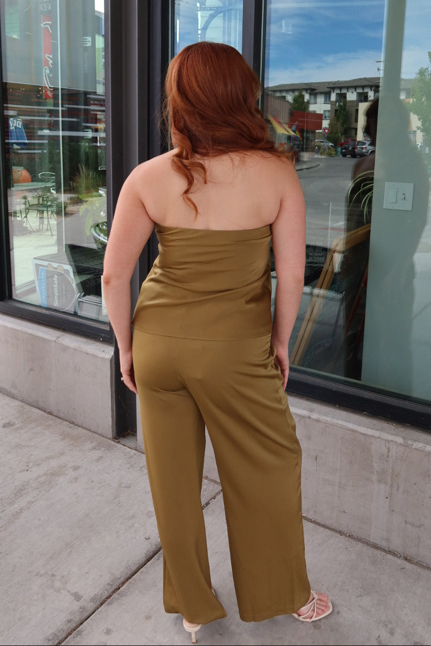 A woman with red hair is seen from behind wearing a strapless olive green jumpsuit while standing on a sidewalk in front of a storefront with large windows.