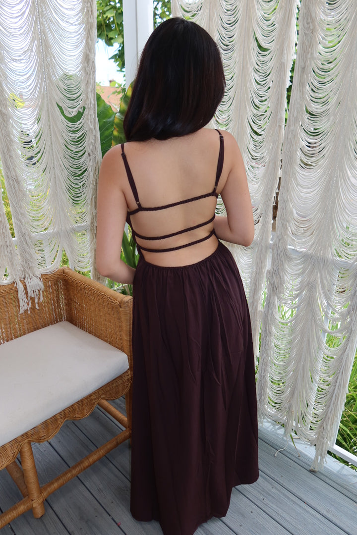 A woman viewed from behind wearing a dark brown backless maxi dress with thin shoulder straps and multiple horizontal strappy details across the upper back. She is standing on a wooden deck in front of white macrame curtains.