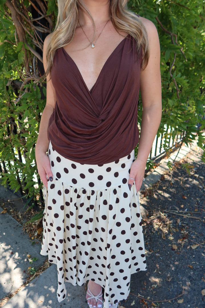 A woman wearing a sleeveless brown wrap-style top and a white skirt with dark brown polka dots, standing outdoors in front of green foliage.