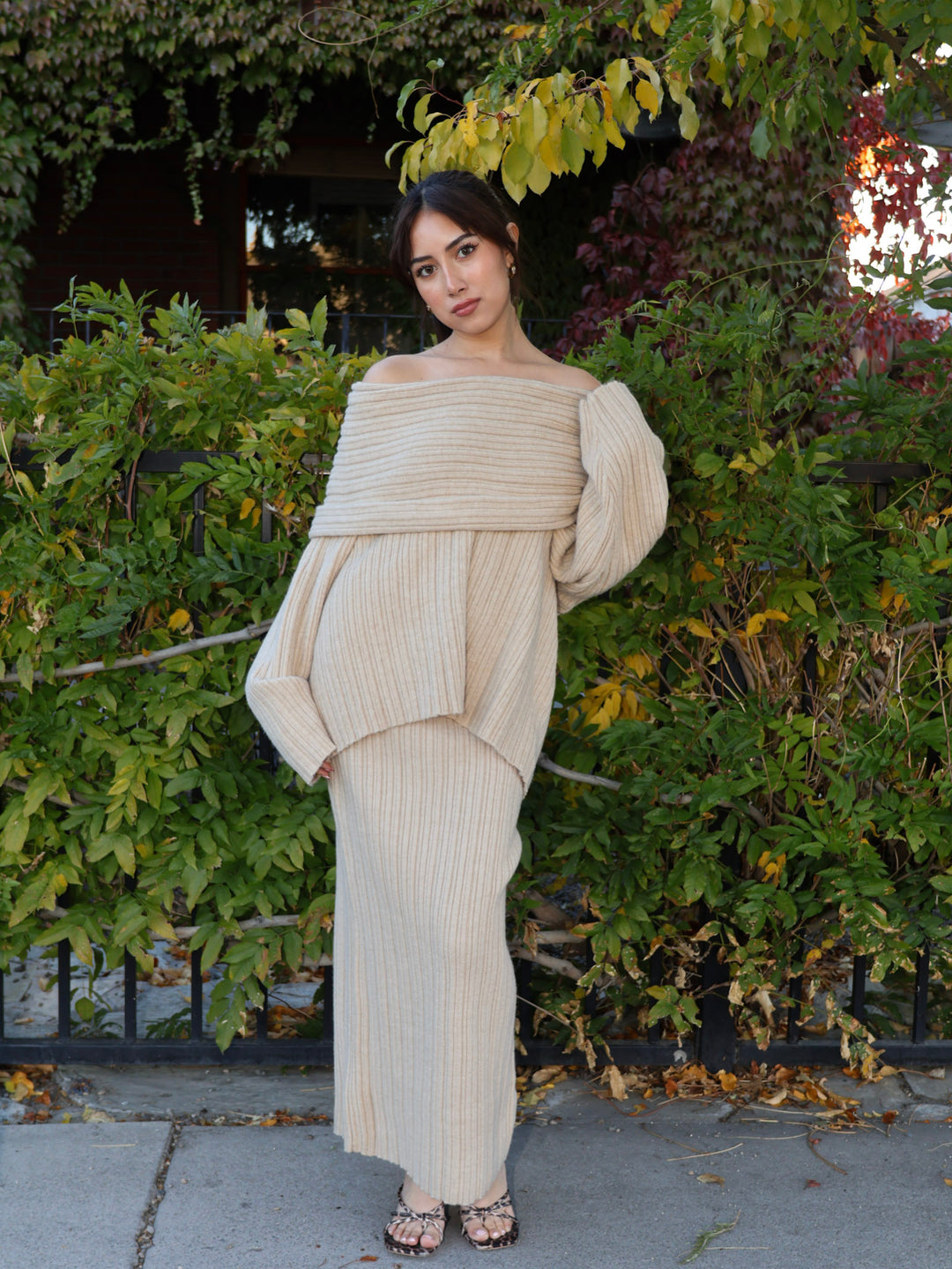 Woman wearing a beige off-shoulder dress standing in front of green bushes.