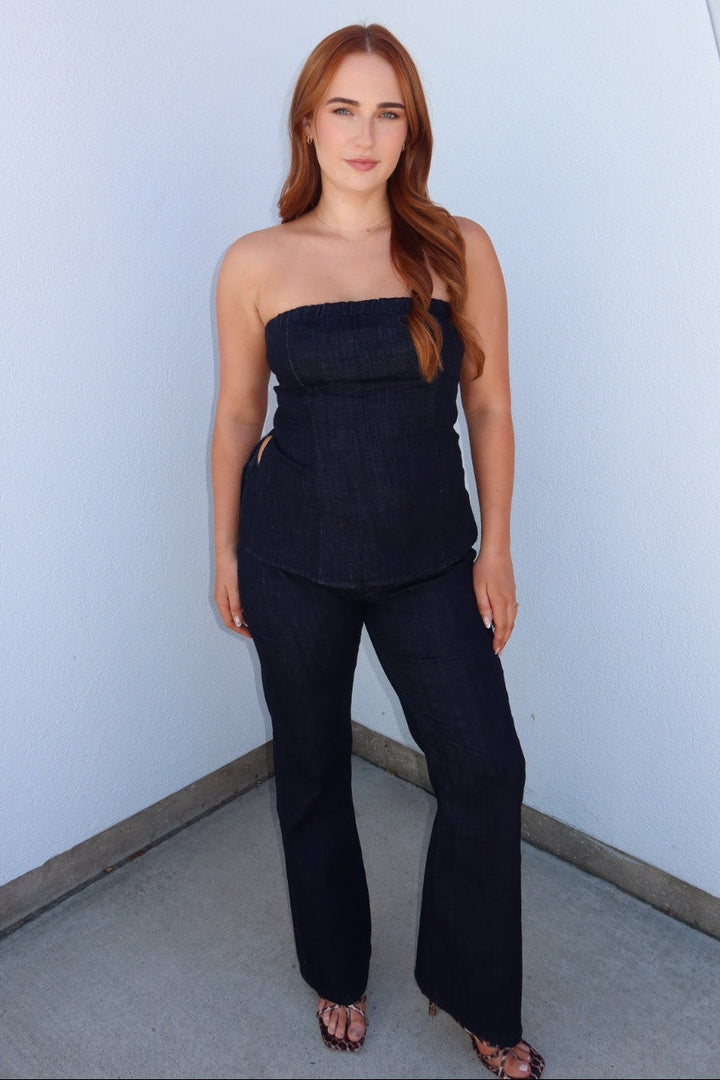 A woman with long red hair stands in a corner wearing a dark navy strapless top and matching dark navy wide-leg pants, paired with brown strappy heels, against a white wall with a red accent.