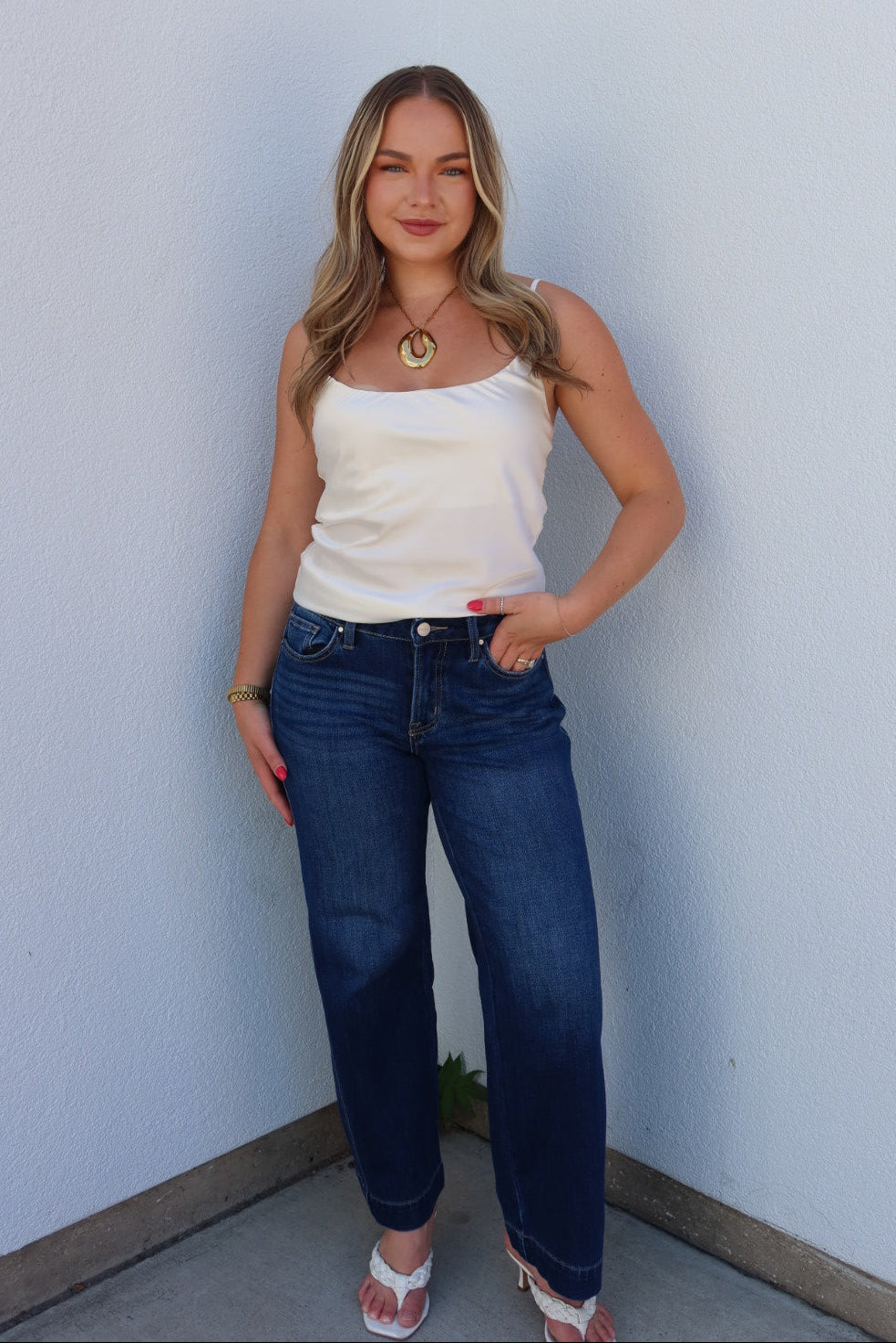 A woman stands in the corner of a light blue wall wearing a white tank top, dark blue jeans, and white slide sandals.