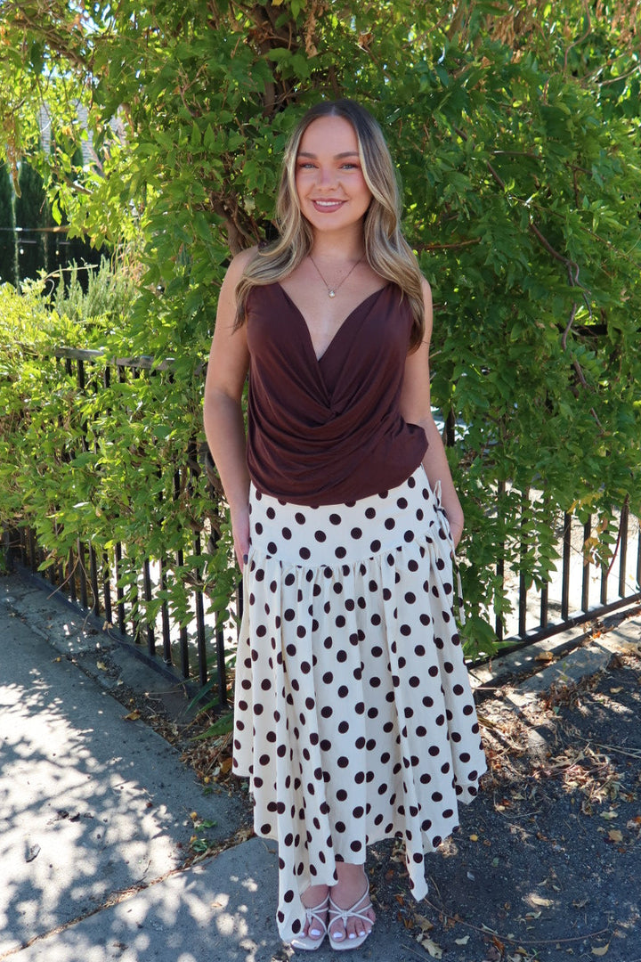 A woman stands outdoors on a sidewalk in front of a leafy green tree, wearing a dark brown draped sleeveless top and a white midi skirt with brown polka dots.