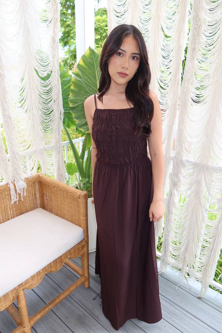A woman modeling a sleeveless, smocked bodice maxi dress in a deep plum-burgundy color, standing on a wooden deck in front of white macrame curtains.