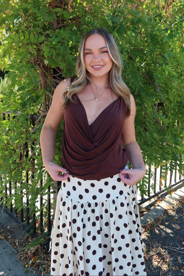 A woman posing outdoors in front of a green hedge and black metal fence, wearing a dark brown draped sleeveless top and a cream skirt with black polka dots.