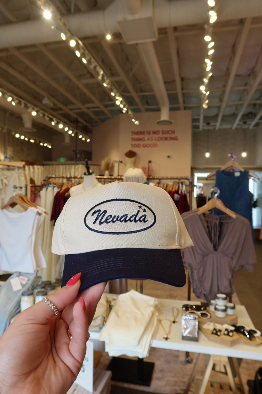 A hand with red nail polish holds up a beige and navy baseball cap featuring an oval 'Nevada' logo on the front, set against a blurred boutique background.