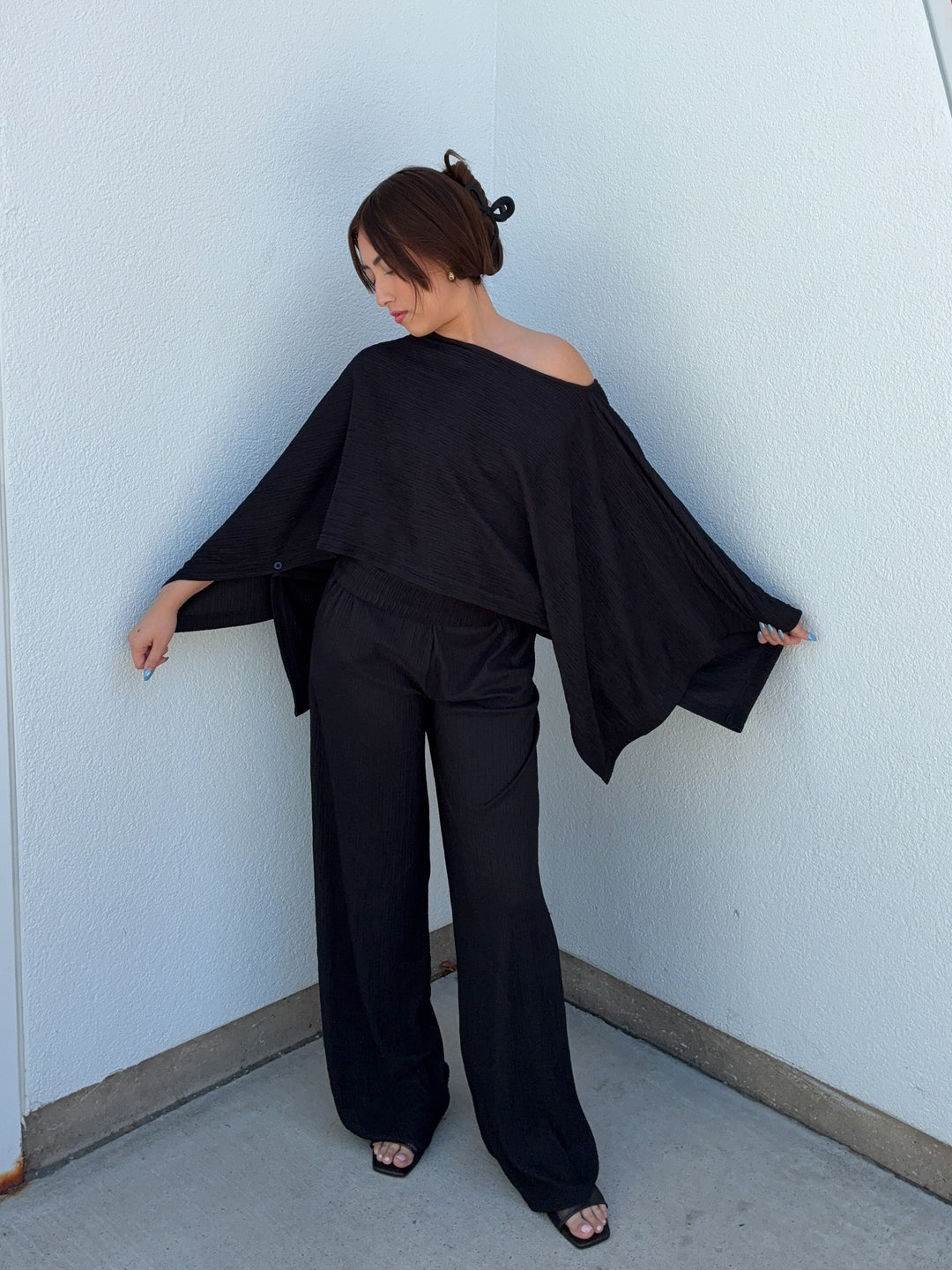 Woman wearing a black off-shoulder top and wide-leg pants against a light blue wall.