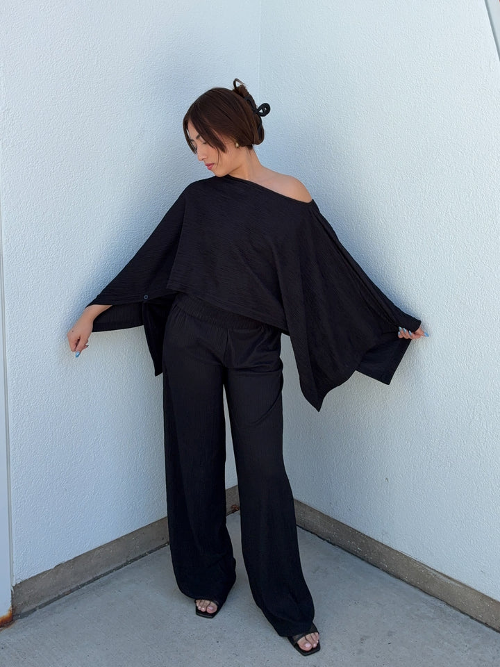 Woman wearing a black off-shoulder top and wide-leg pants against a light blue wall.