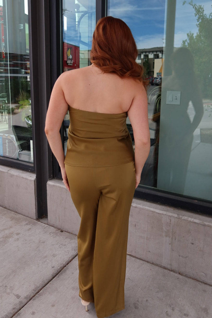 A woman with red hair is shown from behind wearing a strapless olive-brown jumpsuit while standing on a sidewalk in front of a building with large glass windows.