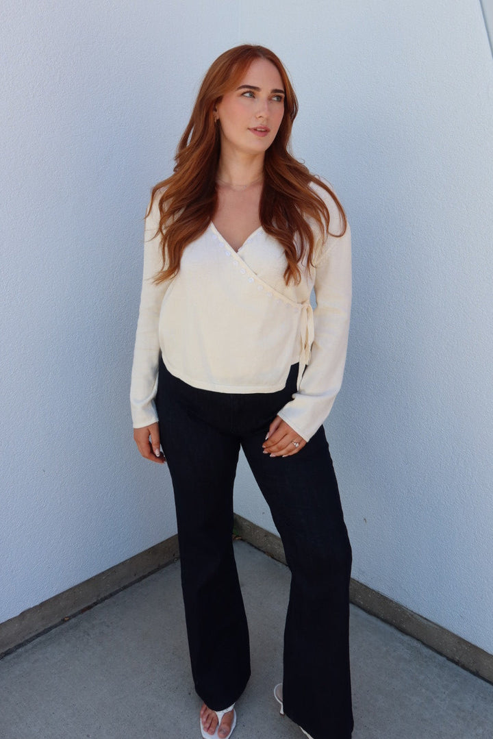 A woman with long red hair stands in a corner wearing a cream wrap-style long-sleeve top, black wide-leg pants, and white sandals against a white and orange wall.