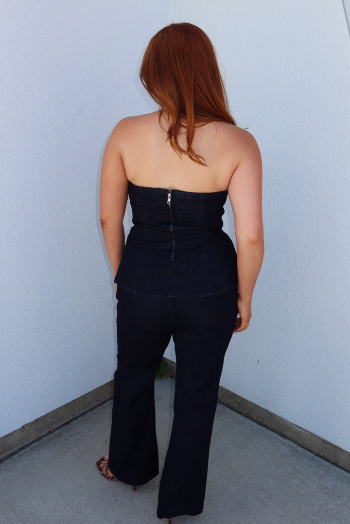A woman with red hair is shown from behind wearing a dark navy strapless denim jumpsuit with a back zipper, standing against a white wall.