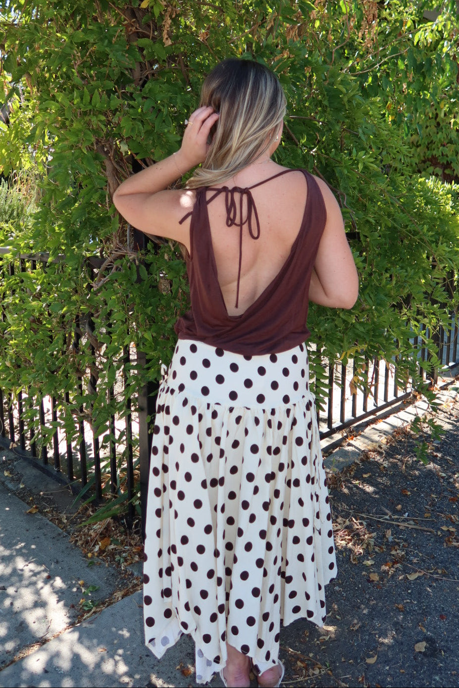 A woman stands outdoors facing away from the camera, wearing a brown open-back top with tie straps and a white skirt with dark brown polka dots. She is positioned in front of a leafy green hedge and a black metal fence.