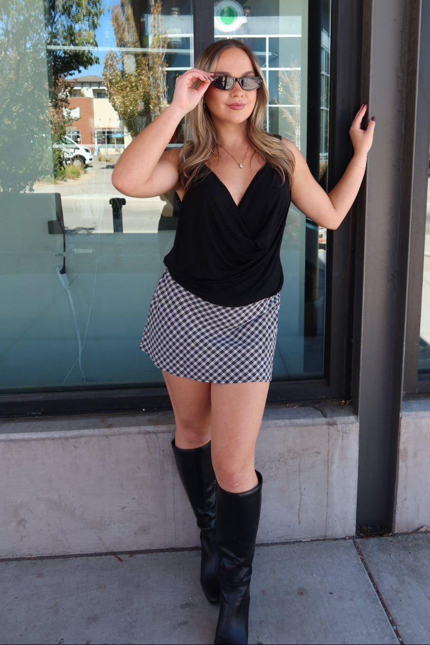 A woman posing outdoors in front of a glass storefront, wearing a black sleeveless top, a black and white checkered mini skirt, and black knee-high boots.