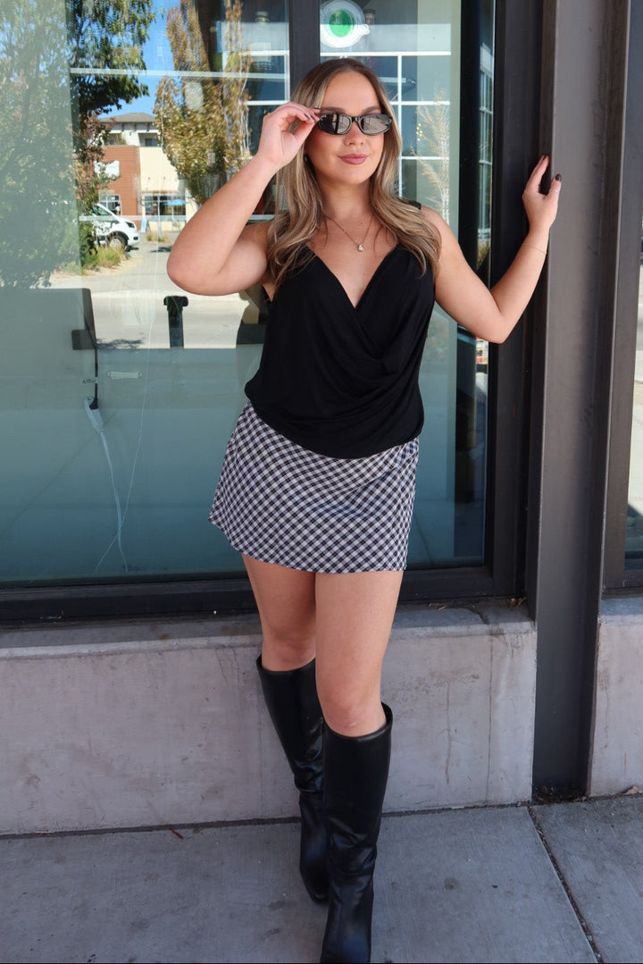 A woman posing outdoors in front of a glass storefront, wearing a black sleeveless top, a black and white checkered mini skirt, and black knee-high boots.