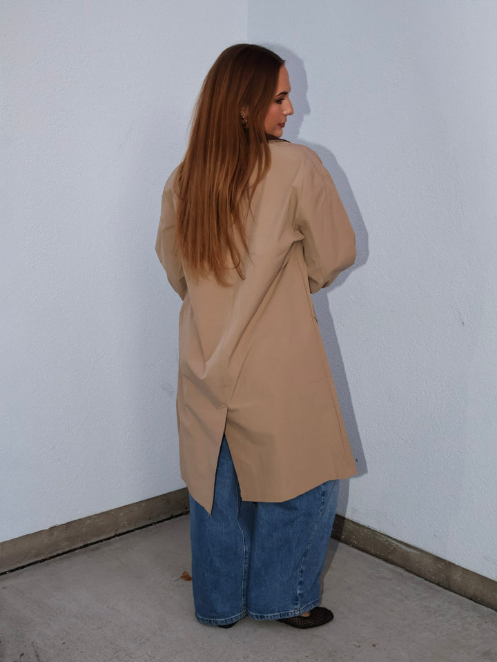 Person wearing a beige coat and blue jeans standing against a white wall.