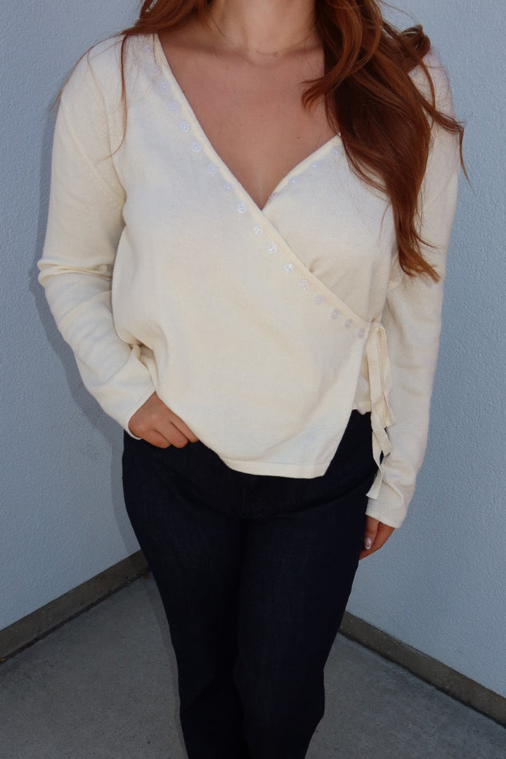 A woman wearing a cream-colored long-sleeve wrap-style top with side buttons and tie details, paired with dark pants and white sandals, standing in a corner against a light gray wall.