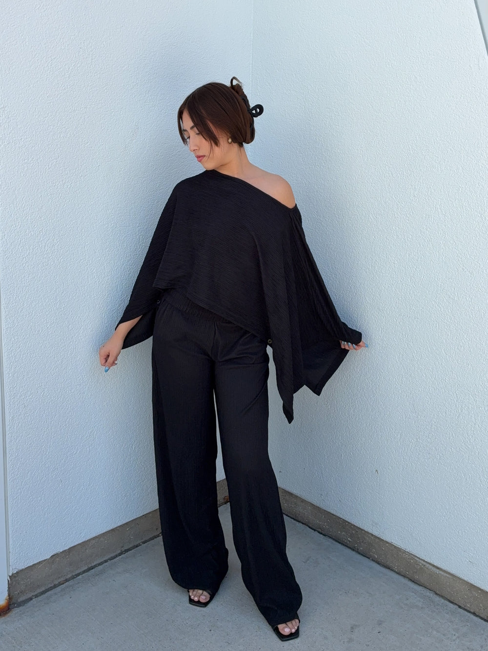 Woman wearing a black off-shoulder top and wide-leg pants against a light blue wall.