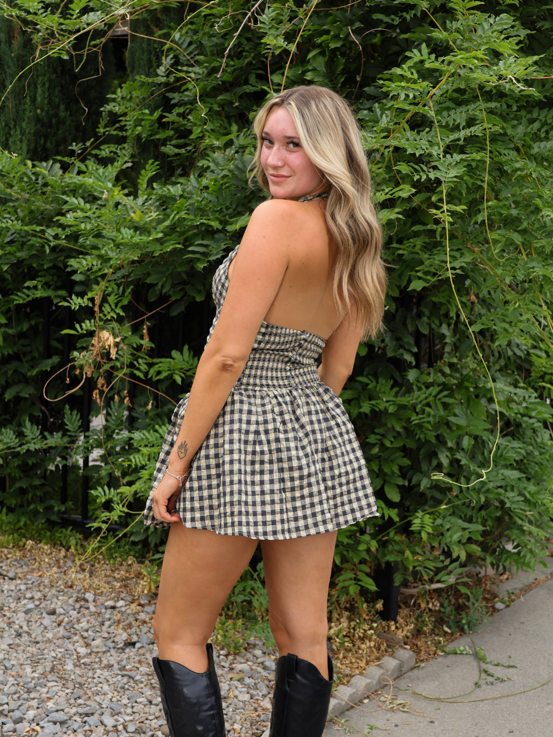 Woman in a checkered dress and black boots standing outdoors with greenery in the background