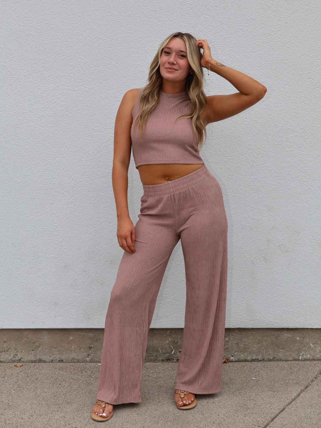 Woman wearing a mauve sleeveless top and wide-leg pants against a white wall.