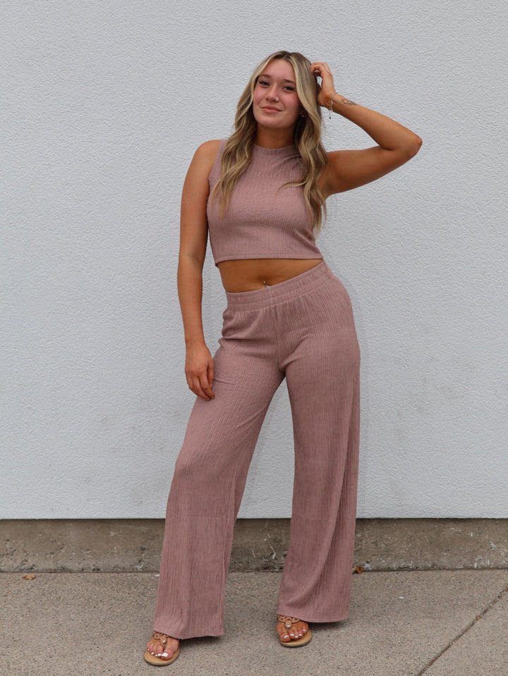 Woman wearing a mauve sleeveless top and wide-leg pants against a white wall.