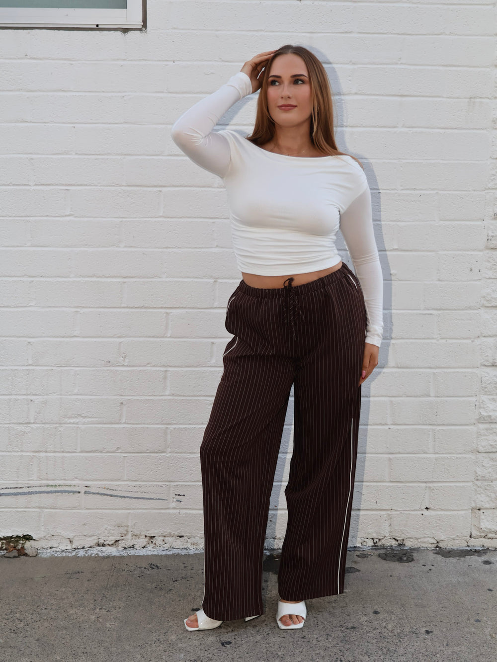 Woman wearing a white long-sleeve top and brown striped pants against a white brick wall.