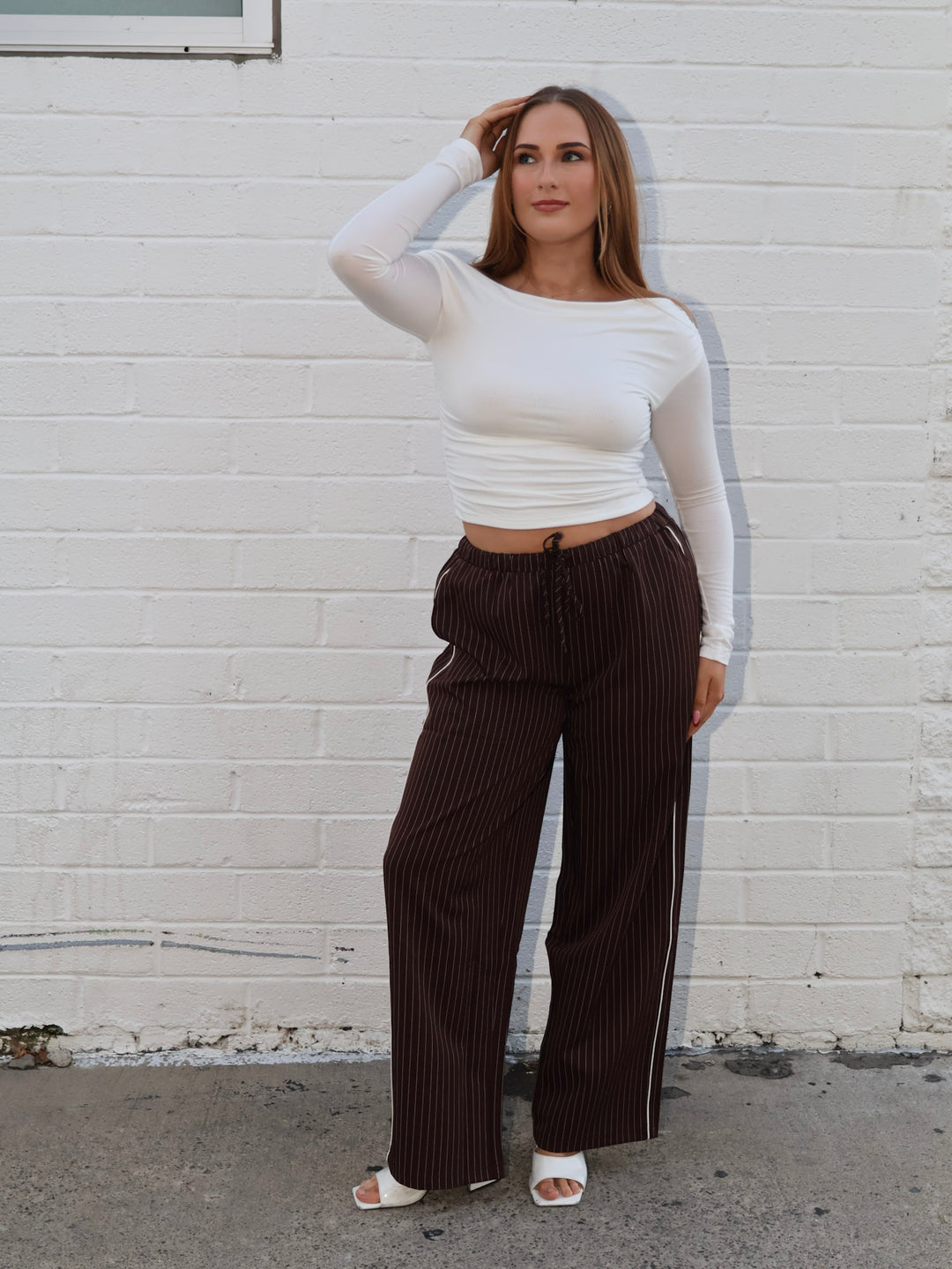 Woman wearing a white long-sleeve top and brown striped pants against a white brick wall.