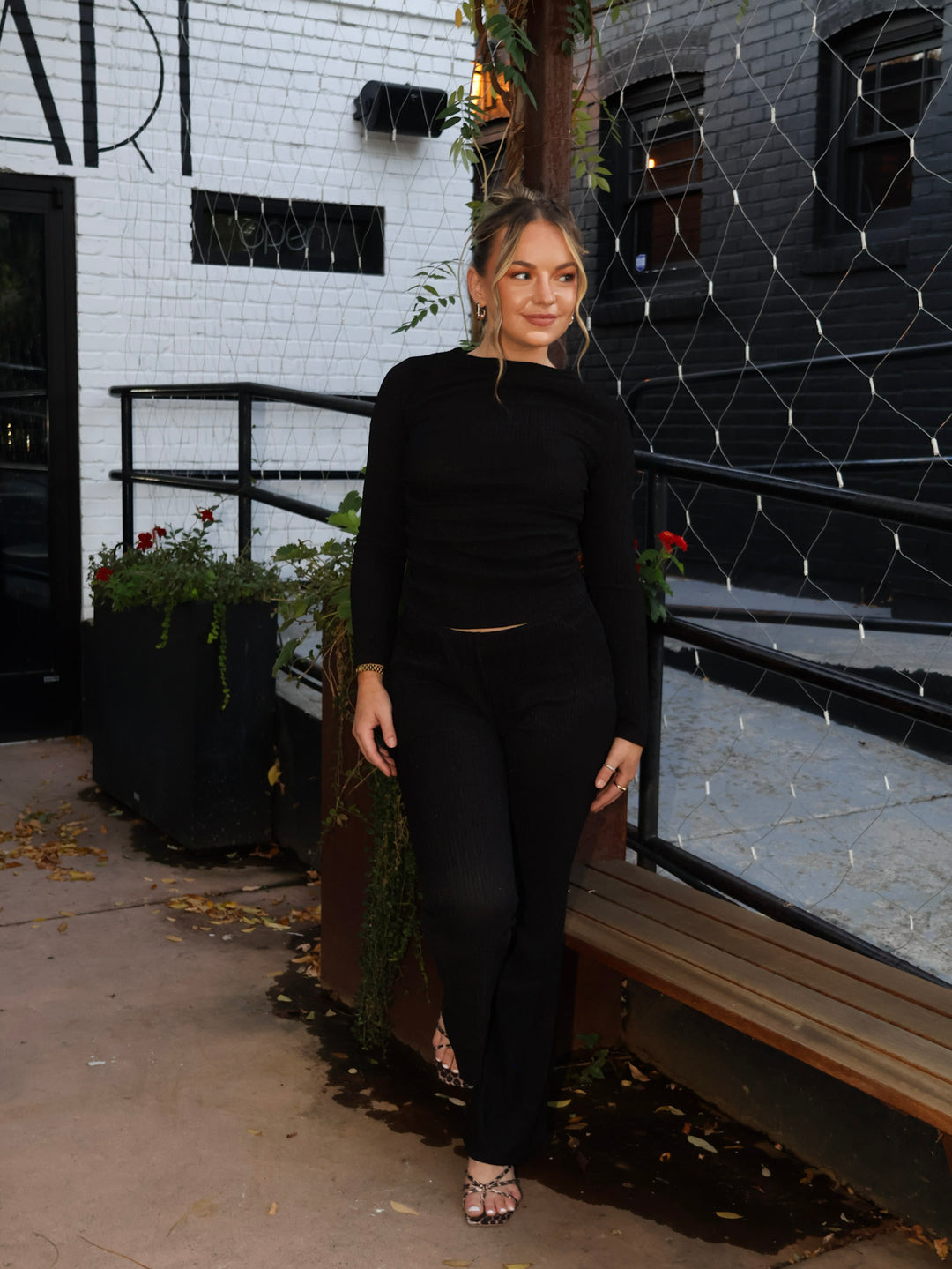 Woman in a black outfit standing in an urban setting with a white brick wall and metal fence.