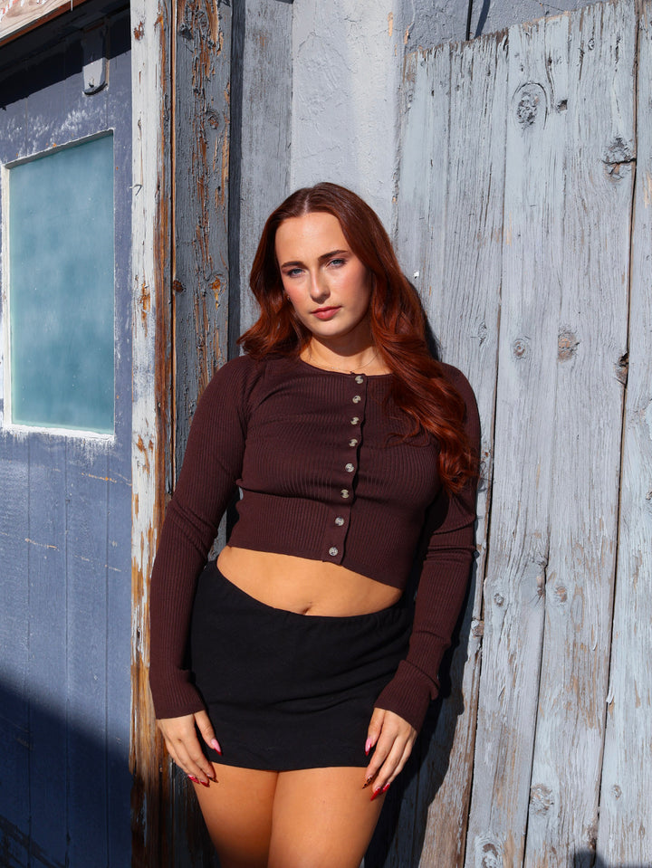 Woman wearing a brown long-sleeve top and black skirt standing against a rustic wooden wall.